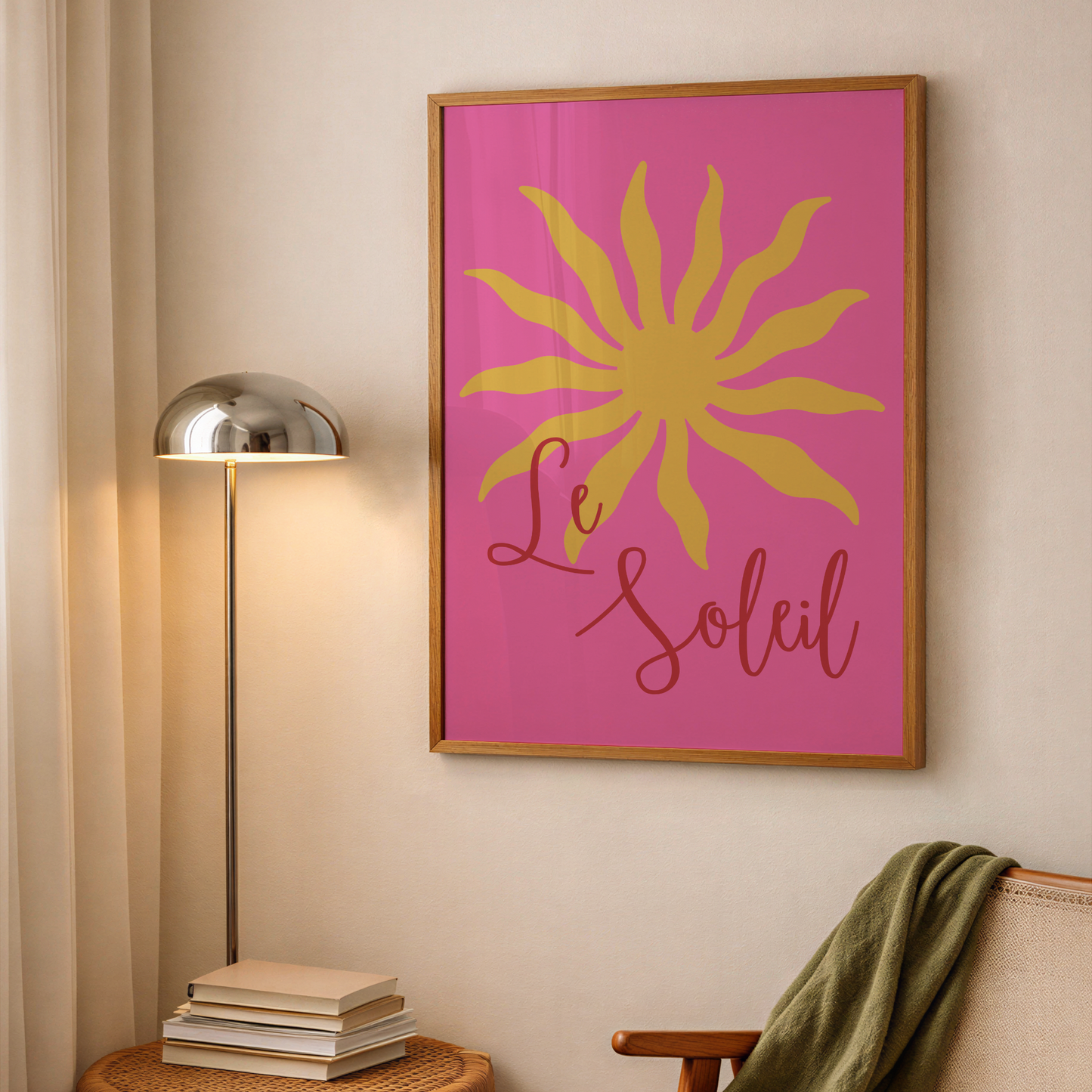 A framed wall art print featuring a yellow sun and the words 'Le Soleil' on a bright pink background, hanging on a beige wall in a styled room with a floor lamp, books, and a chair.