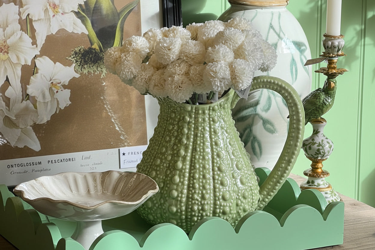 Decorative setting with a green vase, white flowers, and a framed picture on a wooden surface.