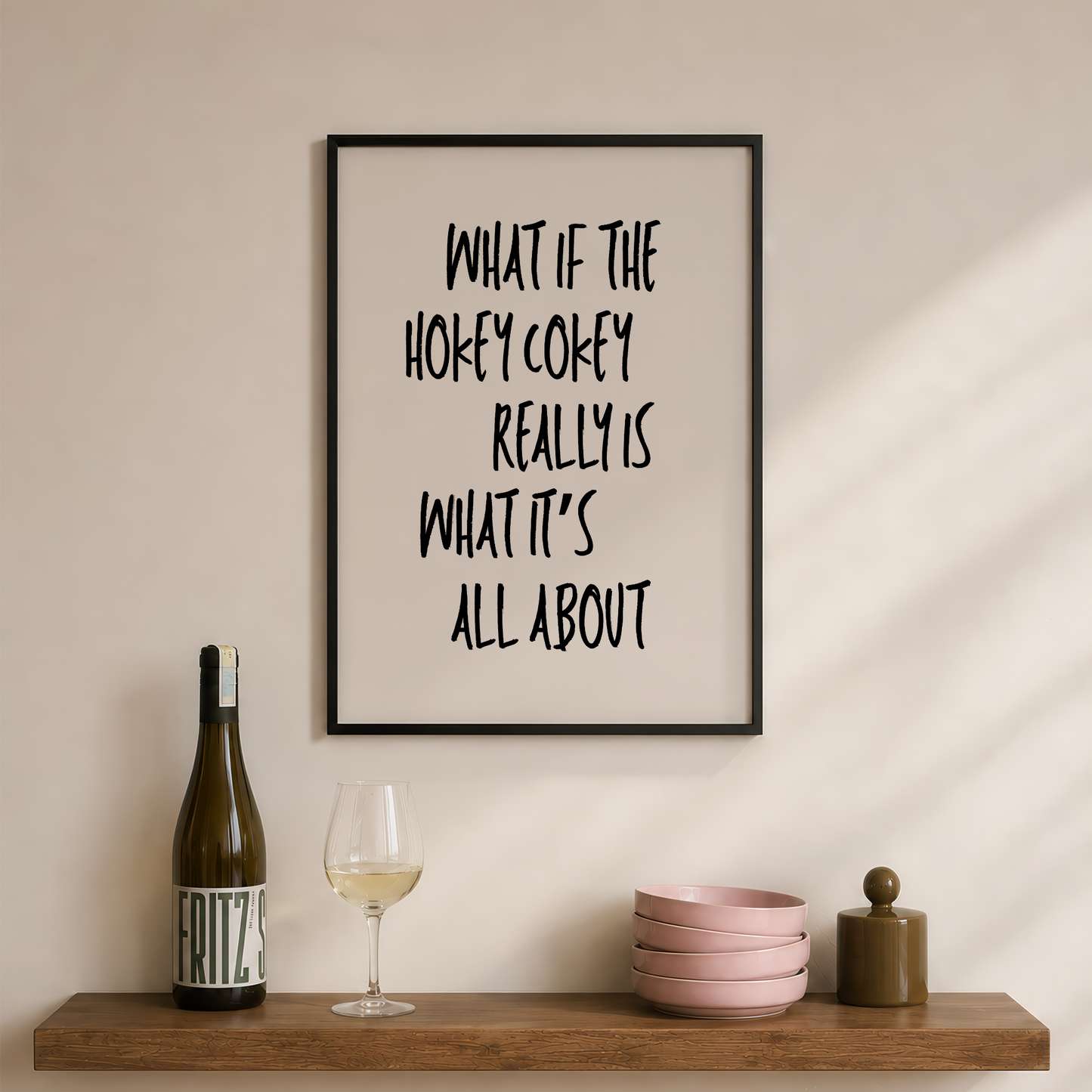 A framed typographic wall print with the phrase “WHAT IF THE HOKEY COKEY REALLY IS WHAT IT’S ALL ABOUT” hangs on a beige wall above a wooden shelf styled with a wine bottle, a glass of white wine, pink bowls, and a small jar.