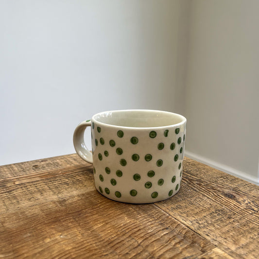 White mug with green polka dots on a wooden surface against a plain wall.