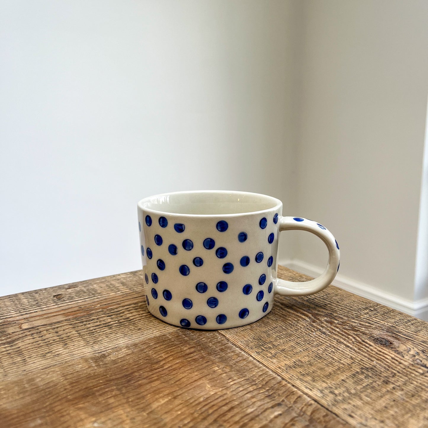 White mug with blue polka dots on a wooden surface against a plain wall.