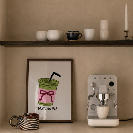 A styled interior scene with a countertop and shelf against a warm beige wall. On the shelf are white and dark blue cups, and on the counter are a framed drink illustration print reading 'MATCHA PLS', a striped mug, and a white SMEG espresso machine.