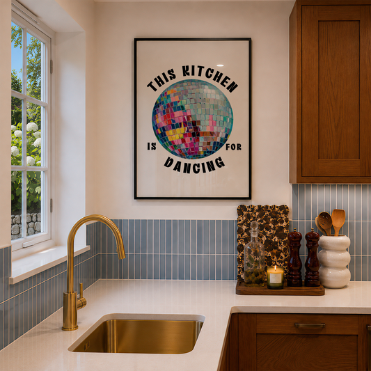 A framed wall art print with the text 'THIS KITCHEN IS FOR DANCING' surrounding a colorful disco ball illustration, hanging in a modern kitchen above a sink and countertop.