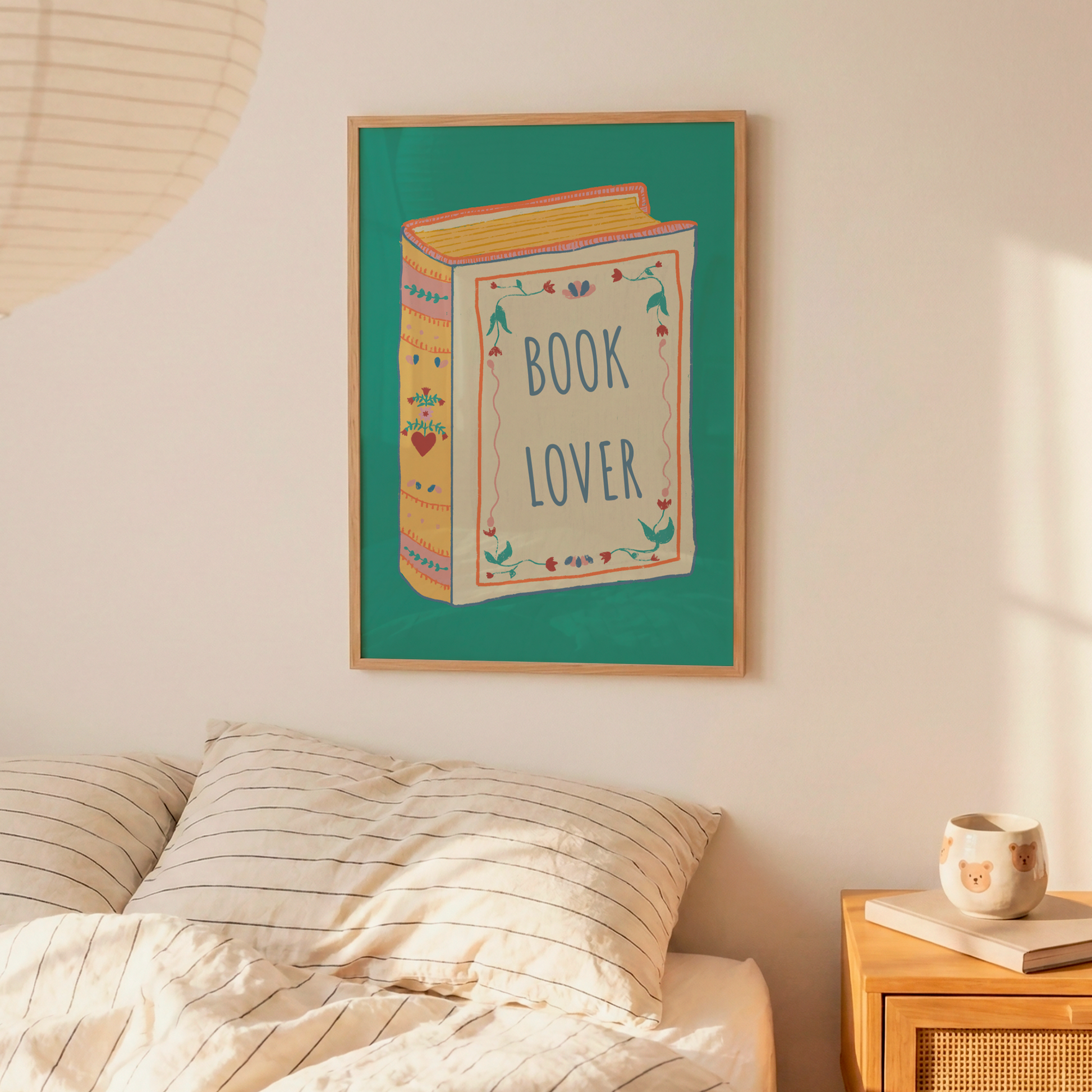 A framed wall art print featuring a colorful illustration of a book with the words 'BOOK LOVER' hangs above a bed in a softly lit bedroom. The scene includes cream bedding, a wooden nightstand with a mug and book, and a warm neutral wall.