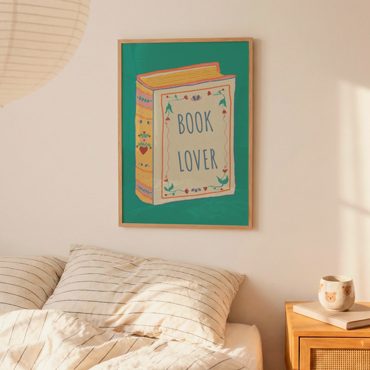 A framed wall art print featuring a colorful illustration of a book with the words 'BOOK LOVER' hangs above a bed in a softly lit bedroom. The scene includes cream bedding, a wooden nightstand with a mug and book, and a warm neutral wall.