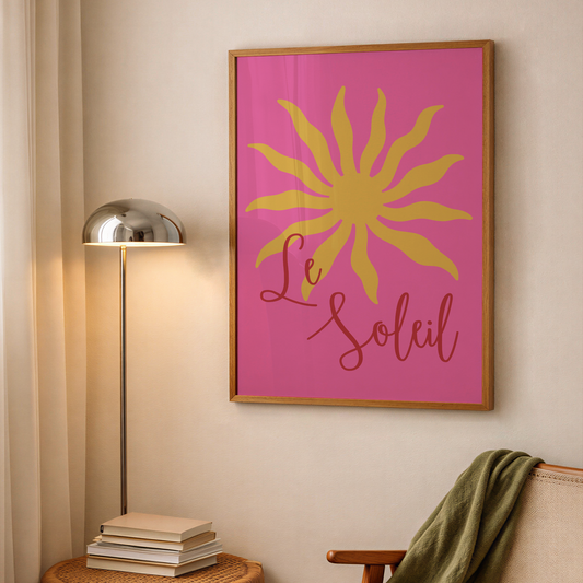 A framed wall art print featuring a yellow sun and the words 'Le Soleil' on a bright pink background, hanging on a beige wall in a styled room with a floor lamp, books, and a chair.