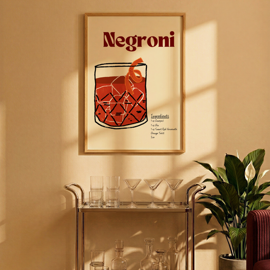 A framed Negroni cocktail poster hangs on a warm beige wall above a glass bar cart with glasses and bottles, next to a potted plant in a sunlit interior.