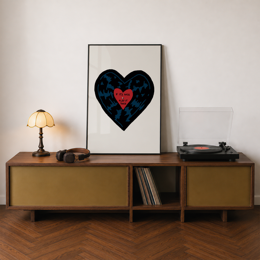 A framed art print featuring a stylized black heart with a red center and the text 'IF IT'S NICE PLAY IT TWICE' is displayed on a wooden console table in a styled room setting with a lamp, headphones, and a turntable.