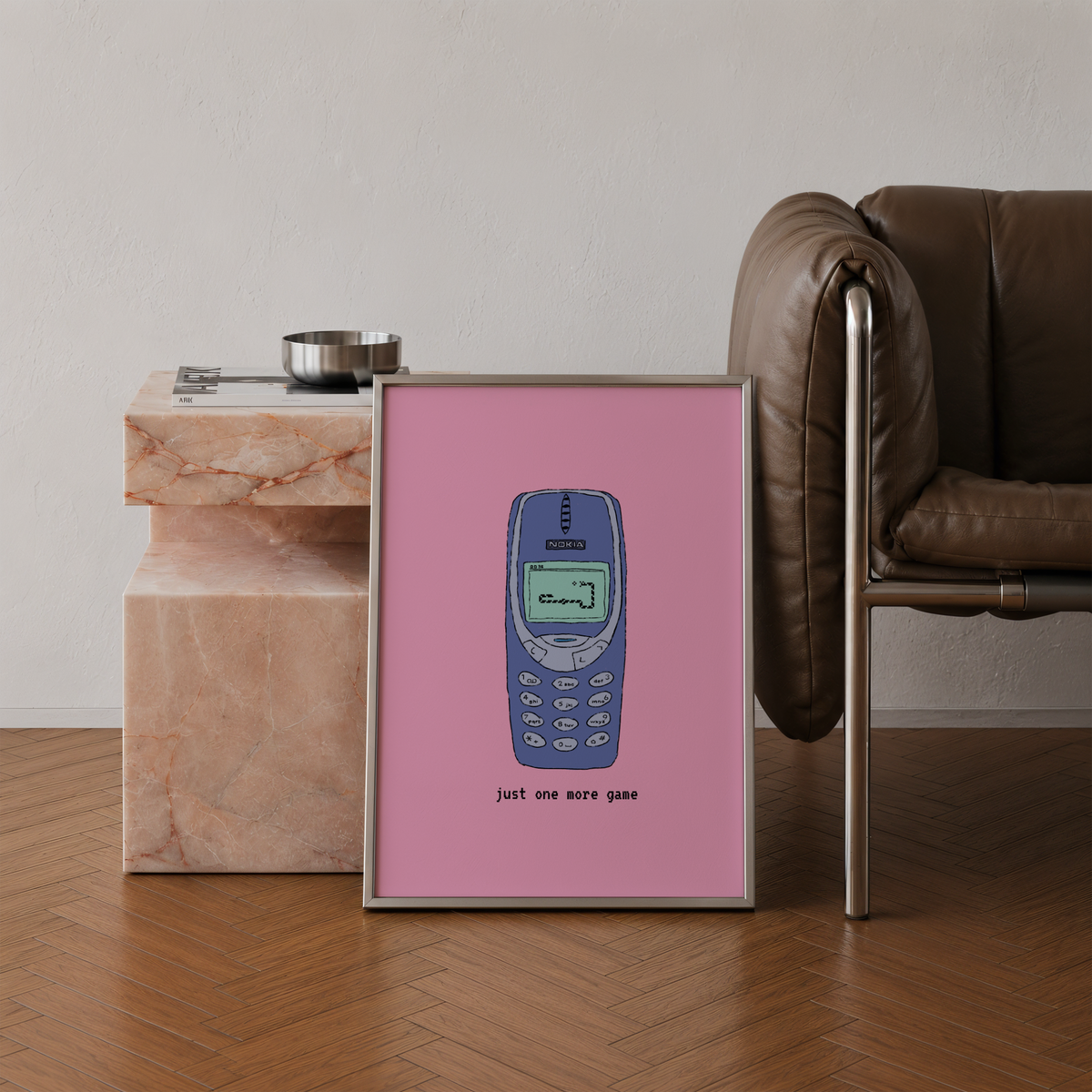 A framed art print featuring a retro blue mobile phone on a pink background, leaning against a marble side table in a modern living room setting with a brown leather chair.