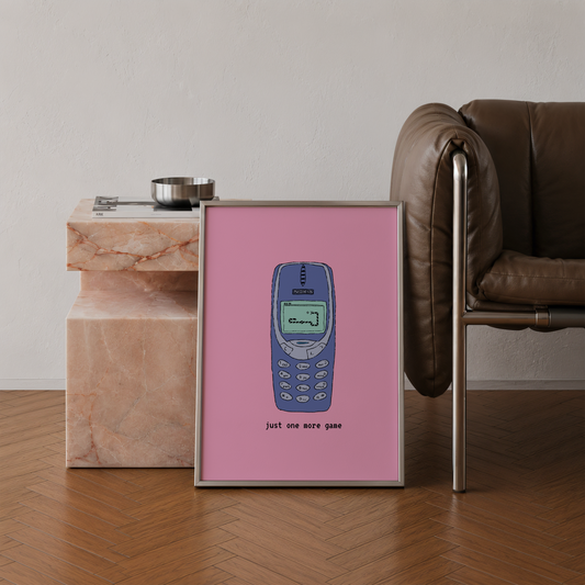 A framed art print featuring a retro blue mobile phone on a pink background, leaning against a marble side table in a modern living room setting with a brown leather chair.